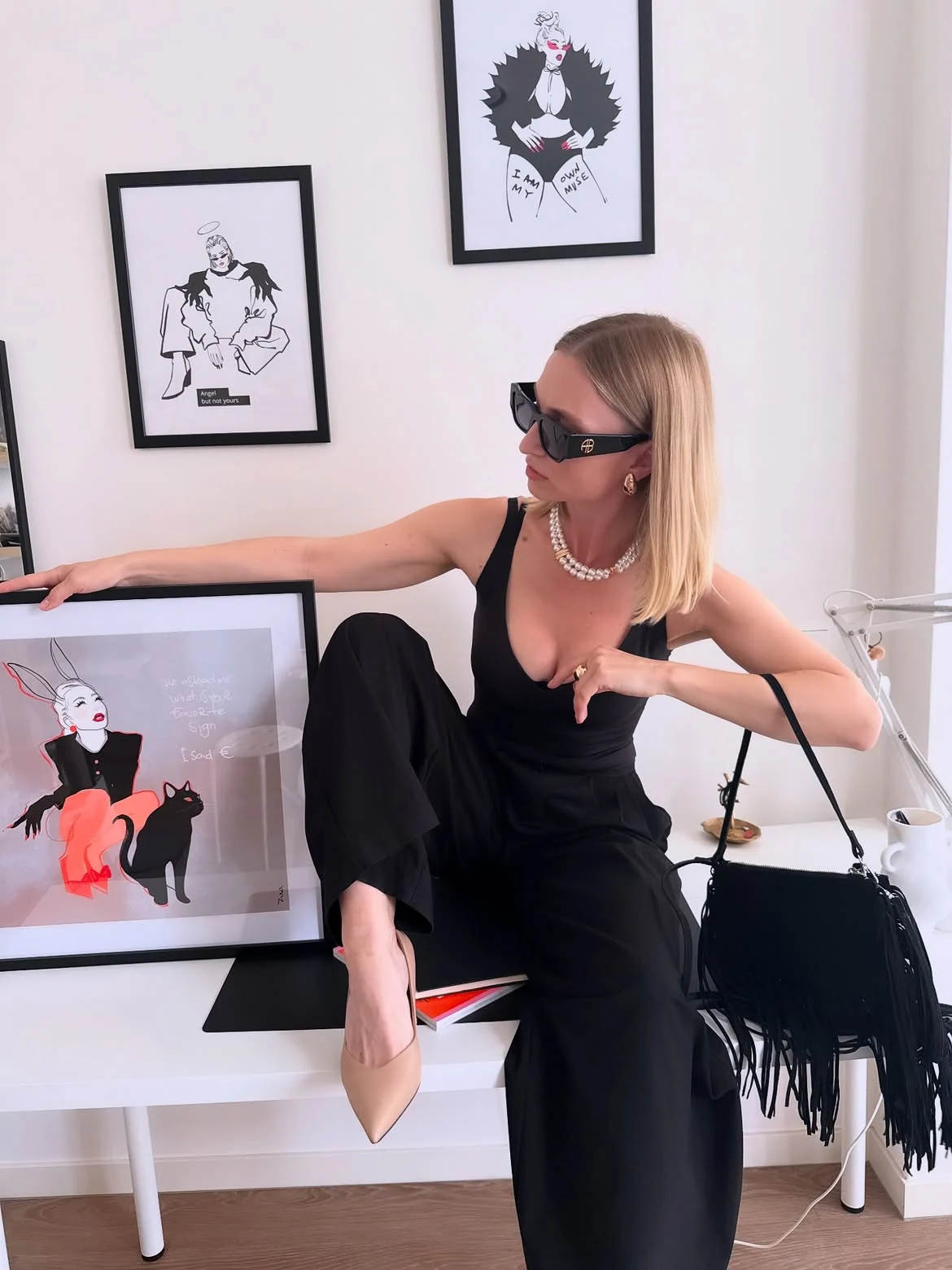 Zina in her studio, seated beside her framed artworks